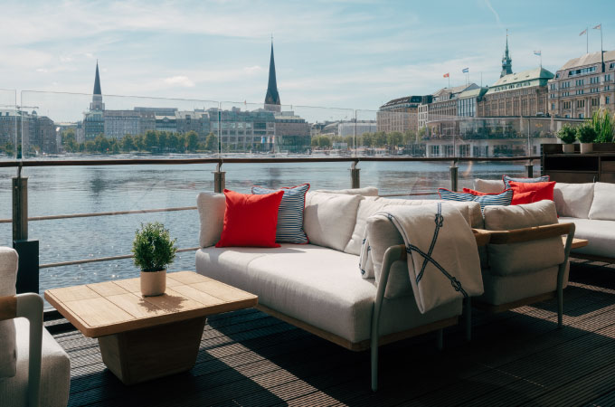 Deck 4 Terrace overlooking a quiet waterfront promenade, furnished with cosy white sofas with red cushions, a small plant on a wooden table and a bright blue sky.