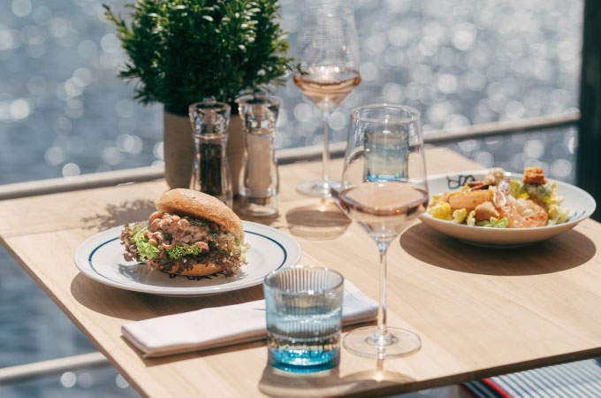 A sunlit table set by the water with a gourmet sandwich, salad, glasses of rose wine, and water.