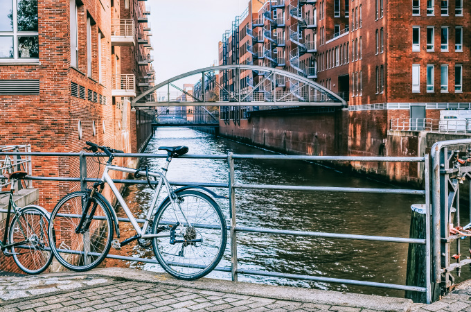 A silver bicycle is attached to a railing overlooking a canal with red brick houses. Water can be seen in the background, with a metal bridge above.