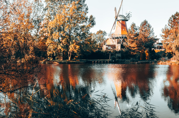 A serene autumn scene with a traditional windmill by a lake. The orange and yellow trees reflect in the water.