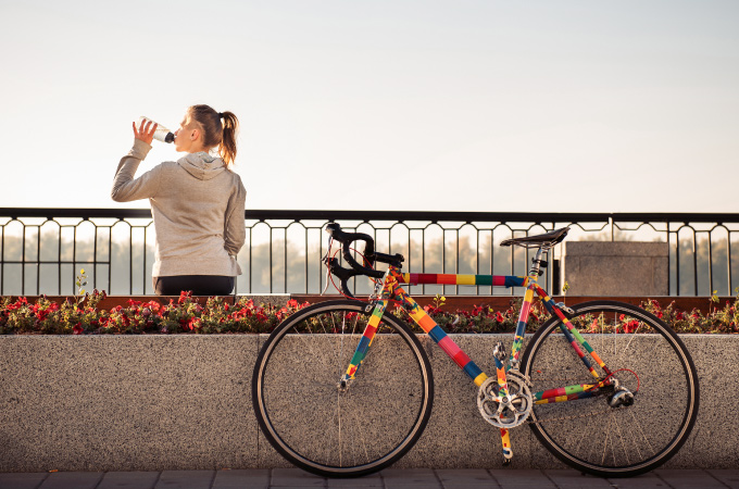 A woman in a gray hoodie drinks water, standing by a railing. Next to her is a colorful, patchwork-patterned bicycle.