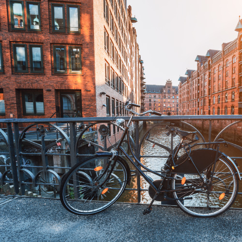 A silver bicycle is attached to a railing overlooking a canal with red brick houses. Water can be seen in the background, with a metal bridge above.
